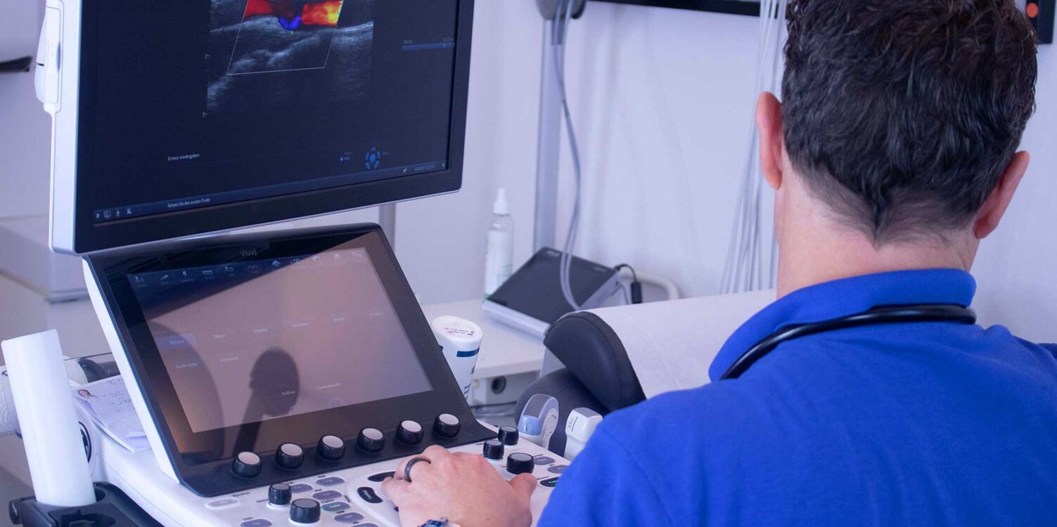 Kardiologie und Angiologie am Lindenplatz Ein Arzt von hinten sitzt an einem Ultraschallgerät in einer Praxis.