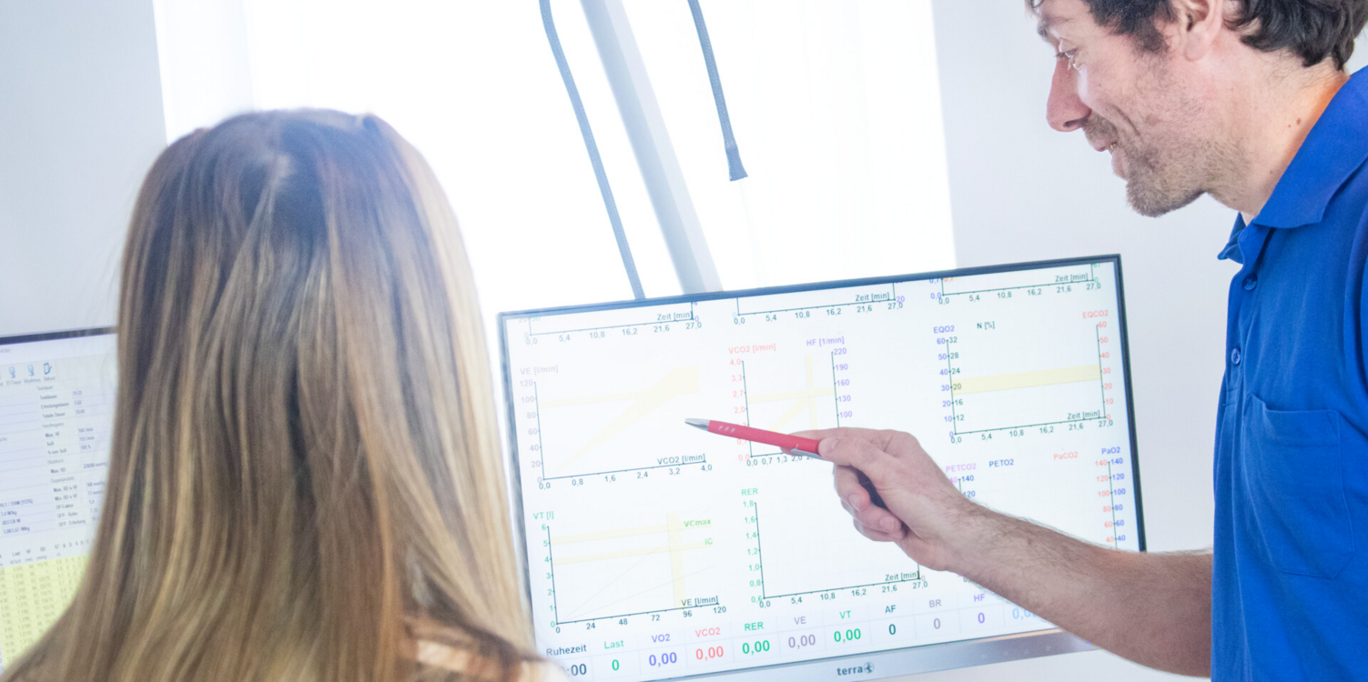 Kardiologie und Angiologie am Lindenplatz - Diabetologie Dr. med. Matthias Mohr von der Seite zeigt mit einem Stift auf einen Bildschirm. Eine junge Patientin mit blonden Haaren von hinten schaut auf den Bildschirm.