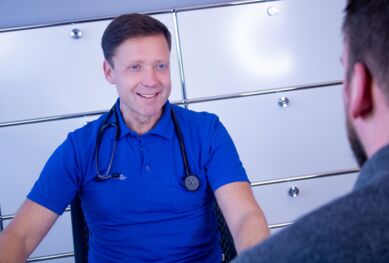 Dr. med.  Langwasser sitzt am Schreibtisch in einem Behandlungszimmer und schaut lächelnd zu einem Patienten, der ihm gegenüber sitzt. Es ist nur ein Teil seines Rücken zu erkennen.